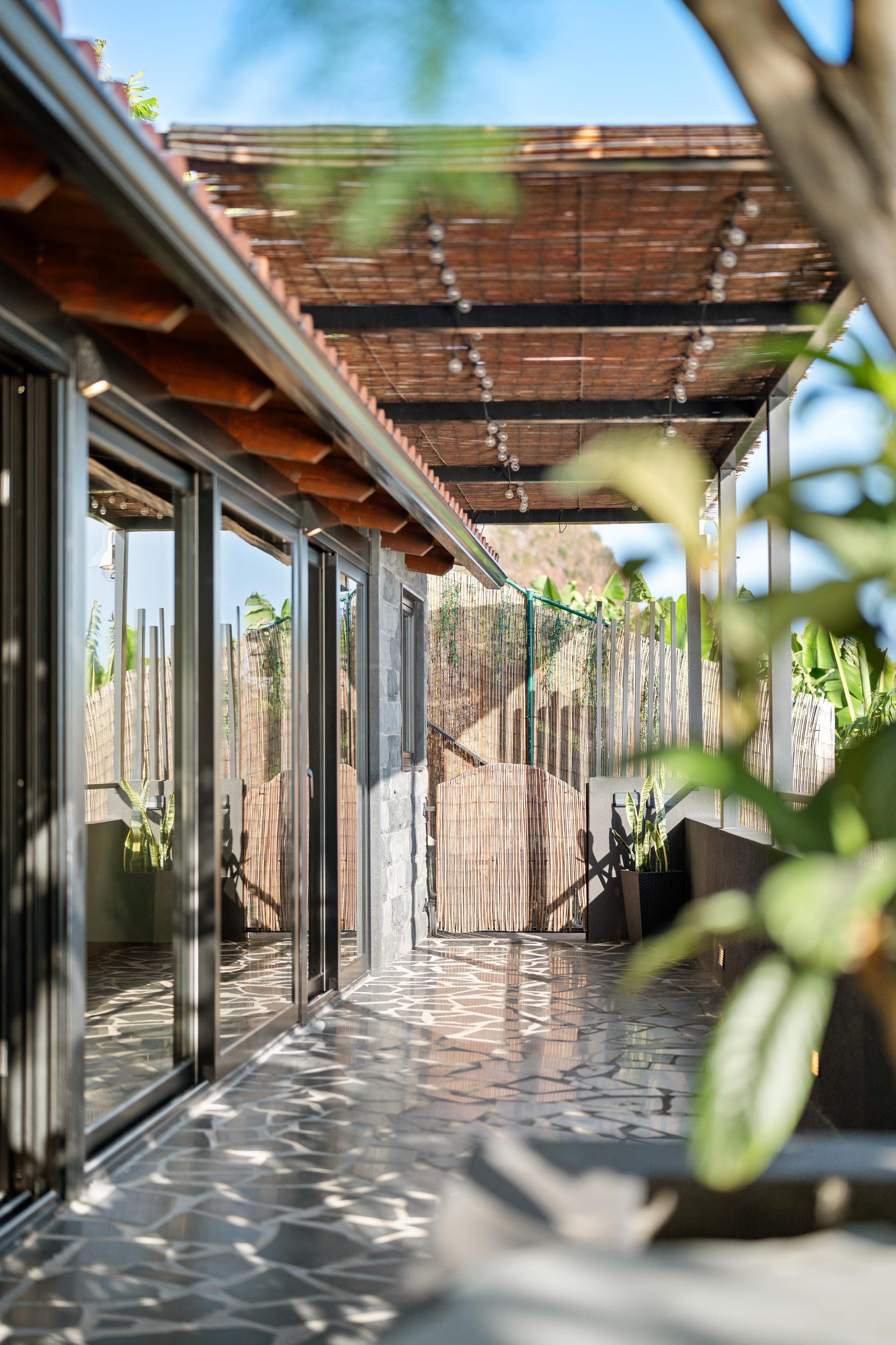The entrance walk in daylight: reed shade overhead, cobblestone underfoot, glass doors along one side.