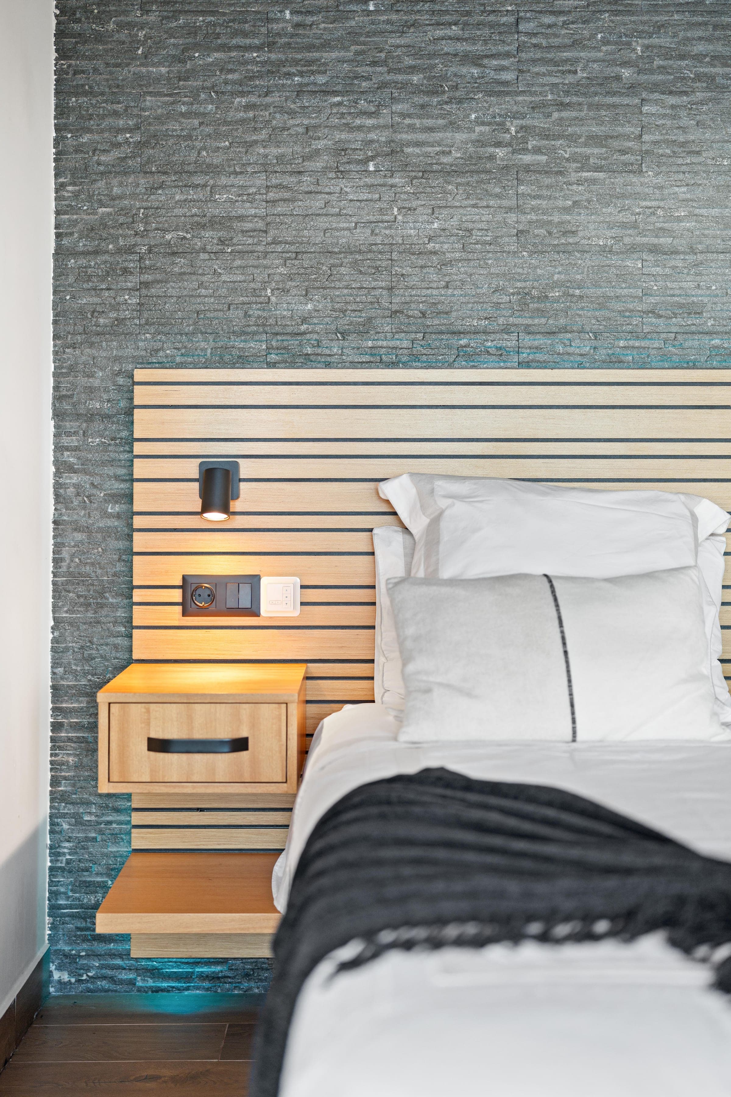 Close up: oak slats, a floating nightstand and a reading light against split stone.