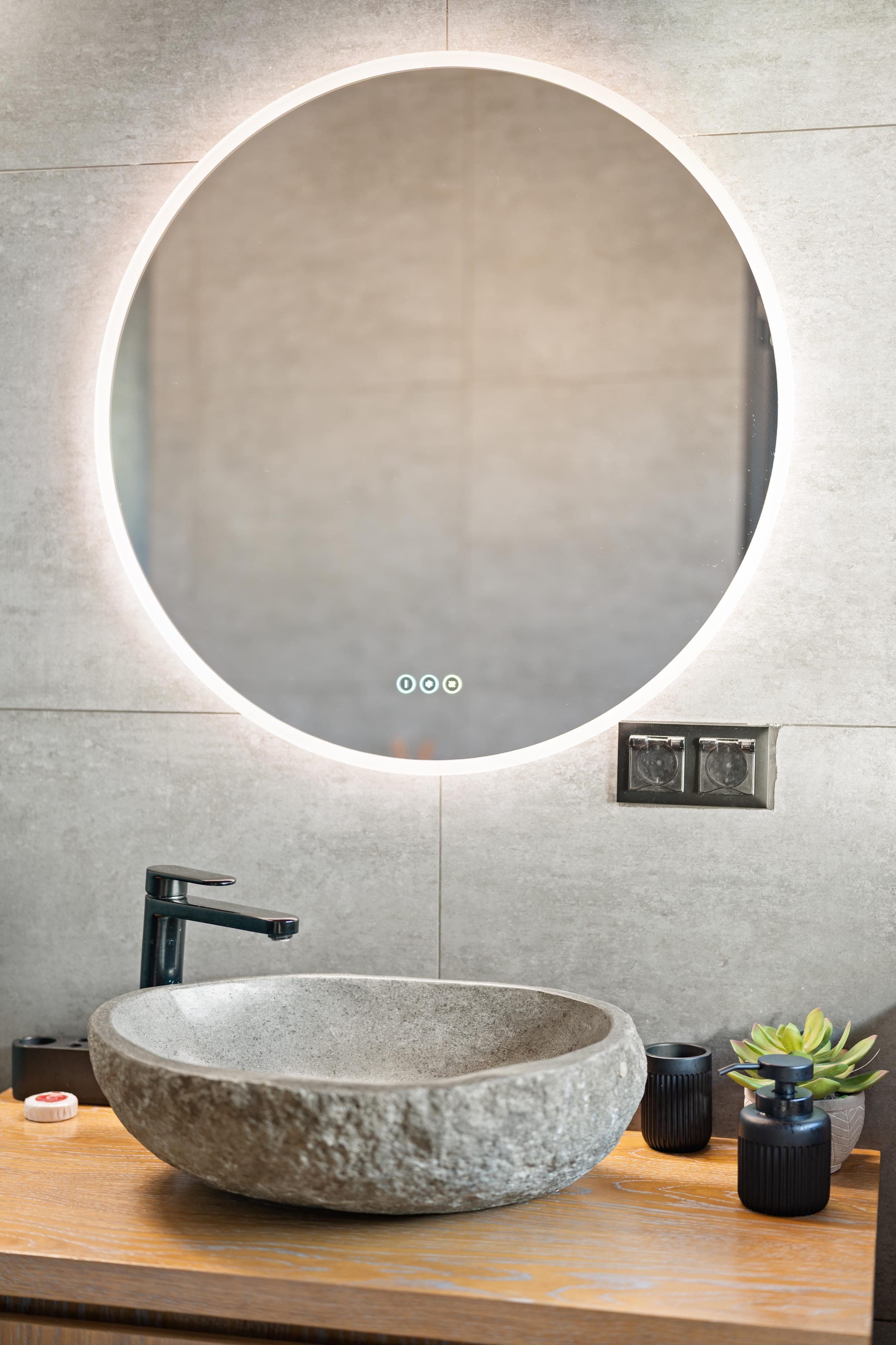 A river-stone basin on oak, under a backlit round mirror.