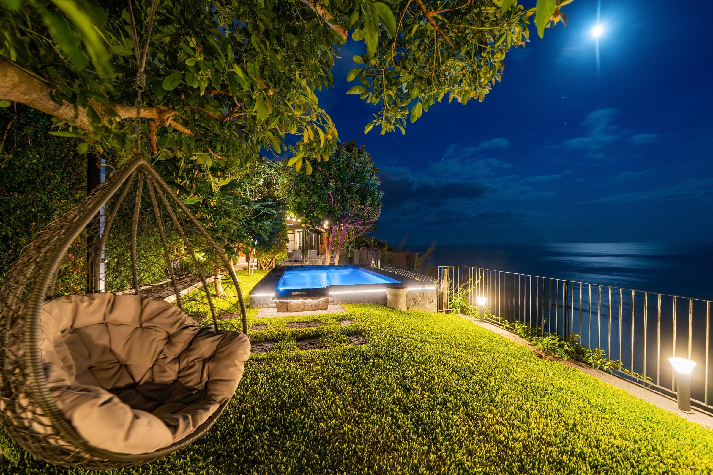 Night: the moon over the water, the pool lit, the hanging chair still.