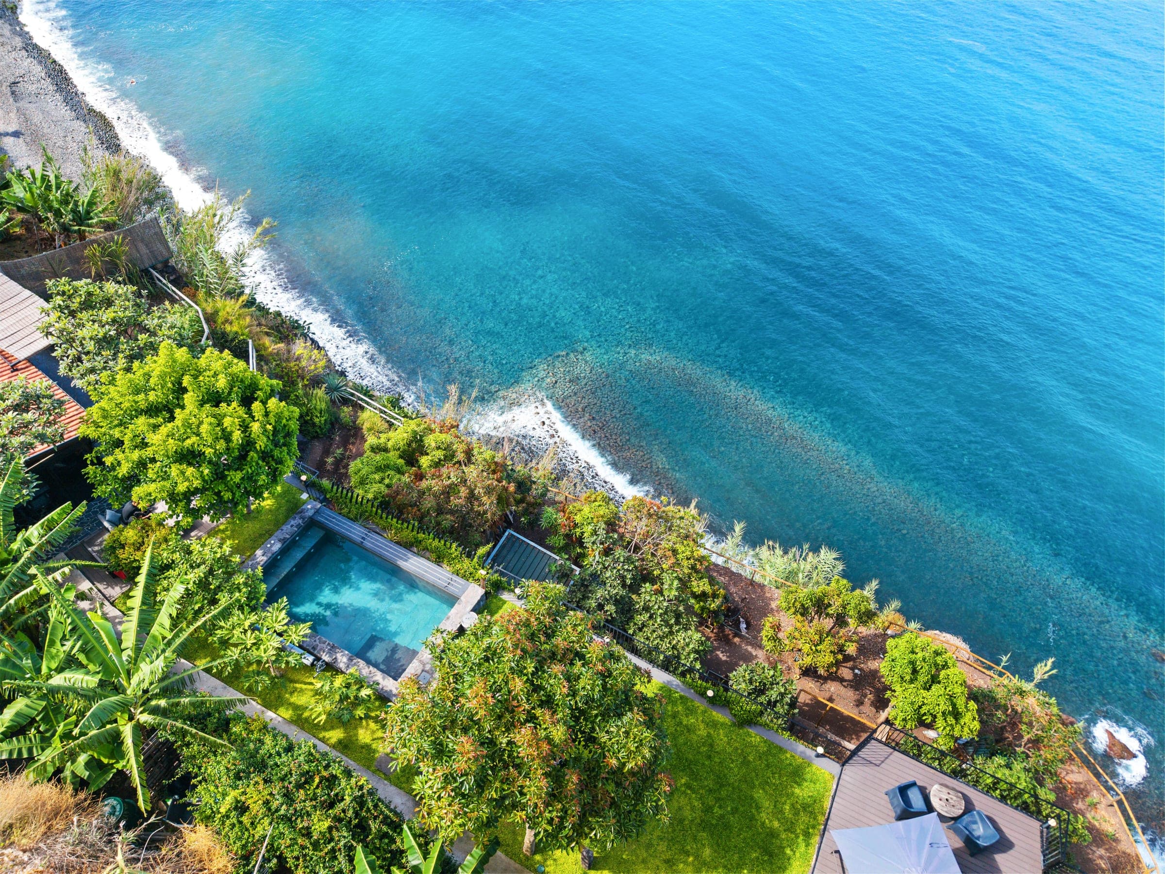 Aerial view of Pomar Escape: an infinity pool between a banana orchard and the black-pebble Atlantic shore.