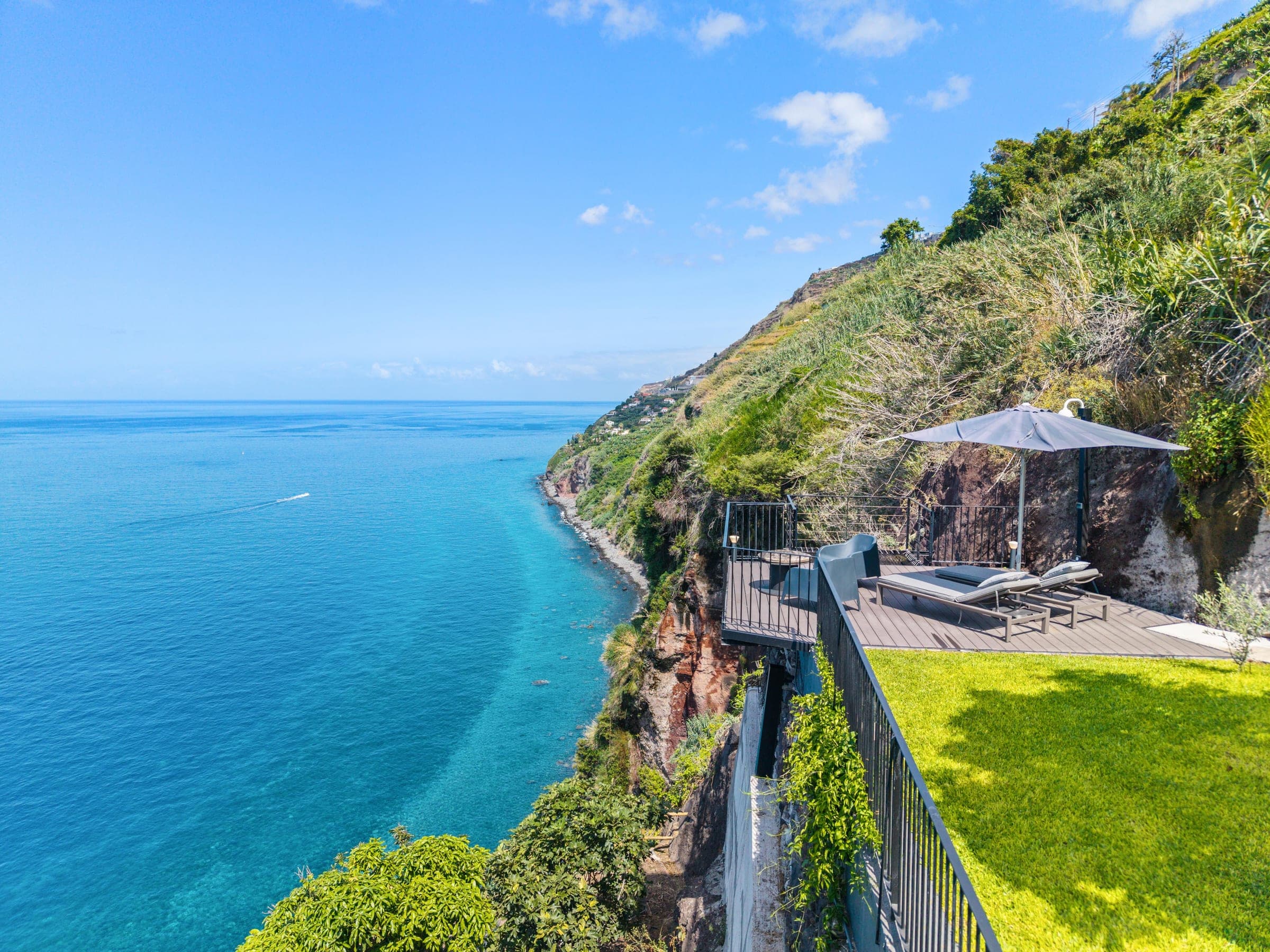 The lawn and the cliff-edge deck from above, turquoise shallows running along the coast.