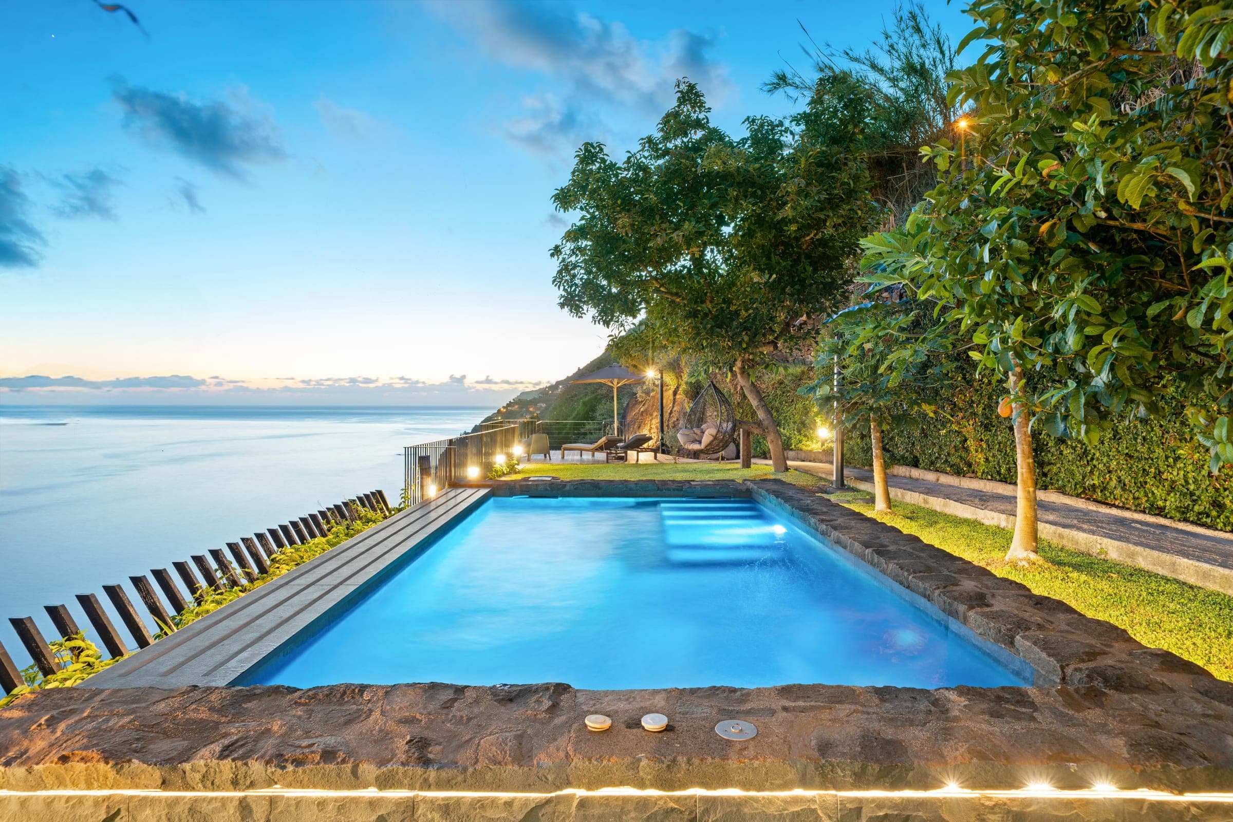 Blue hour at the pool: the infinity edge lit from below, a hanging chair under the tree, and the Atlantic beyond.